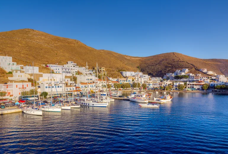 Sailboats and fishing boats moored stern-to along an island harbour quay beneath arid hills