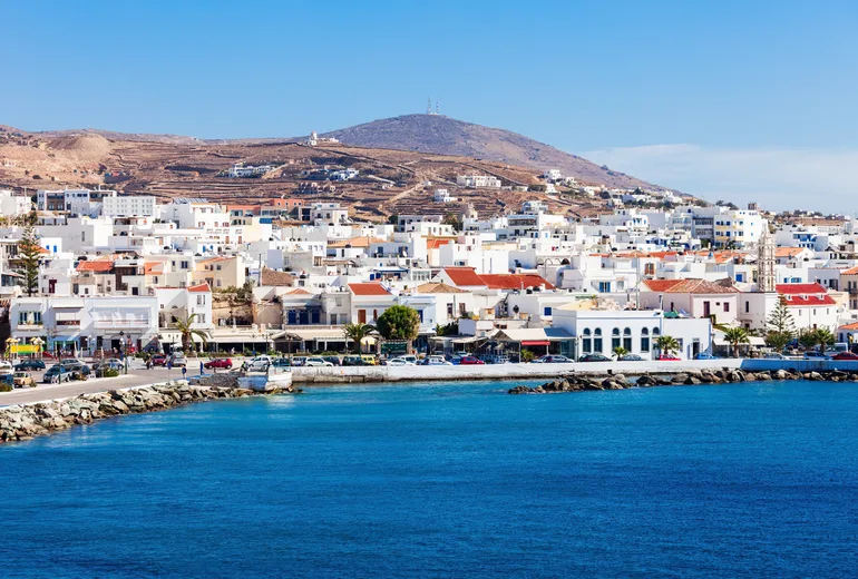 Tinos town waterfront with white buildings and a coastal road beneath bare hills