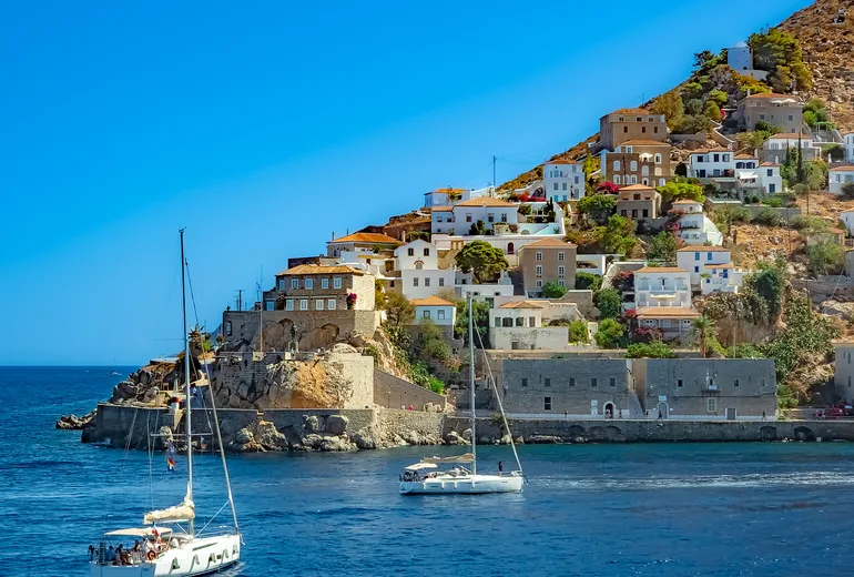Two sailing yachts entering Hydra island harbour below stone hillside houses